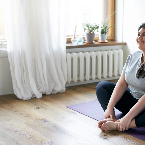 Cheerful attractive young overweight woman in activewear choosing healthy lifestyle, sitting on mat with hands on bare feet, doing butterfly yoga exercise, stretching thighs. Body shape and activity